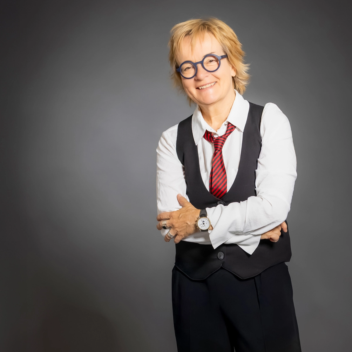 Claudia Puzik ist 60 Jahre alt und etwa 162 cm gross. Sie hat kurze, blonde Haare, lässigen Haarschnitt, sie trägt eine runde, dunkelblaue Brille und lächelt offen und ruhig. Bekleidet ist sie mit einem weissen Hemd, einer schwarzen Weste, einer rot-schwarz gestreiften Krawatte und einer schwarzen Hose; an der Hand trägt sie einen Ring, am Handgelenk eine klassische Armbanduhr mit rundem Gehäuse, hellem Zifferblatt und Lederarmband. Ihre Arme sind locker vor dem Körper verschränkt, die Haltung wirkt aufrecht, selbstbewusst und gelassen, vor einem neutralen Studio-Hintergrund. Sie lächelt.